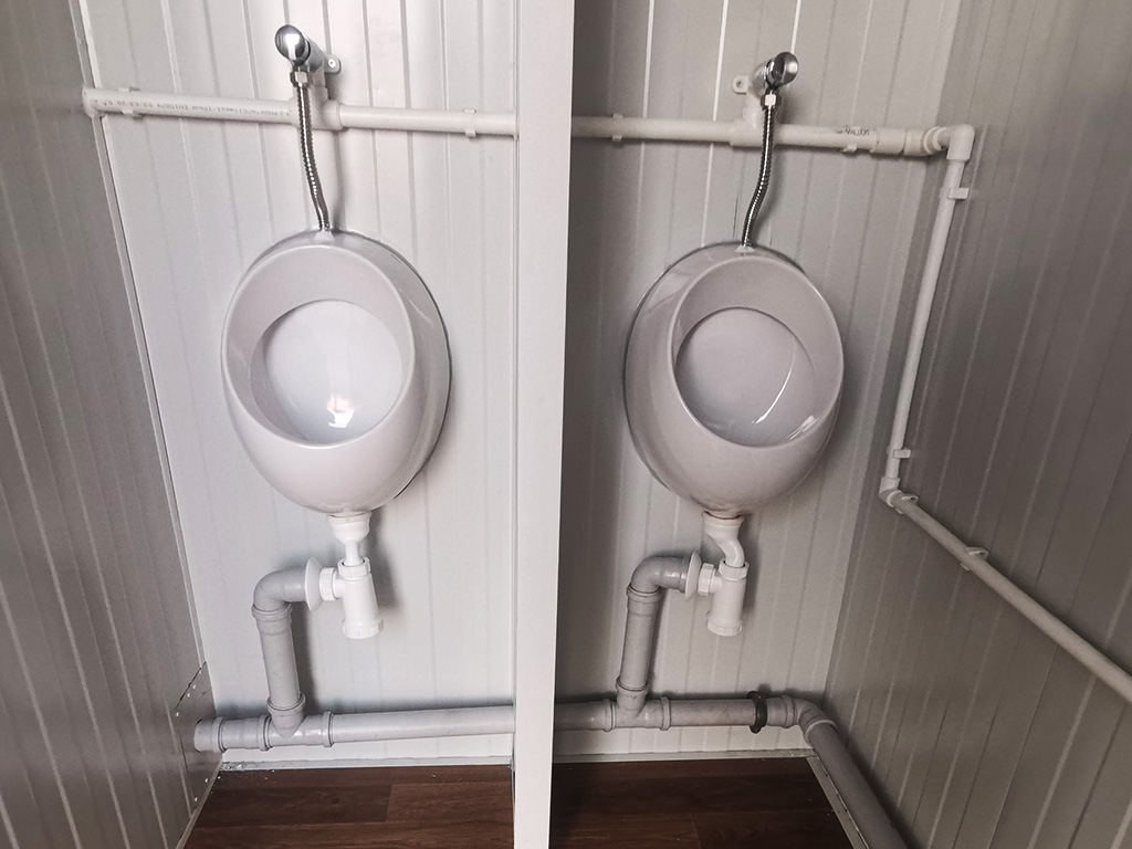 Zwei weiße Urinale nebeneinander in einem Container mit sichtbaren Wasserleitungen