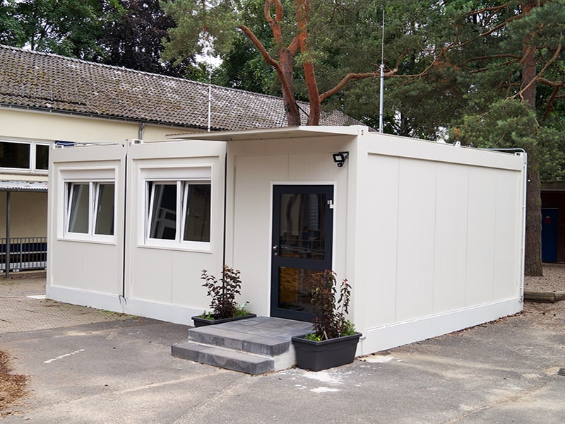 Weißer Bürocontainer mit Fenstern und Eingangstür auf einem befestigten Gelände, umgeben von Bäumen.